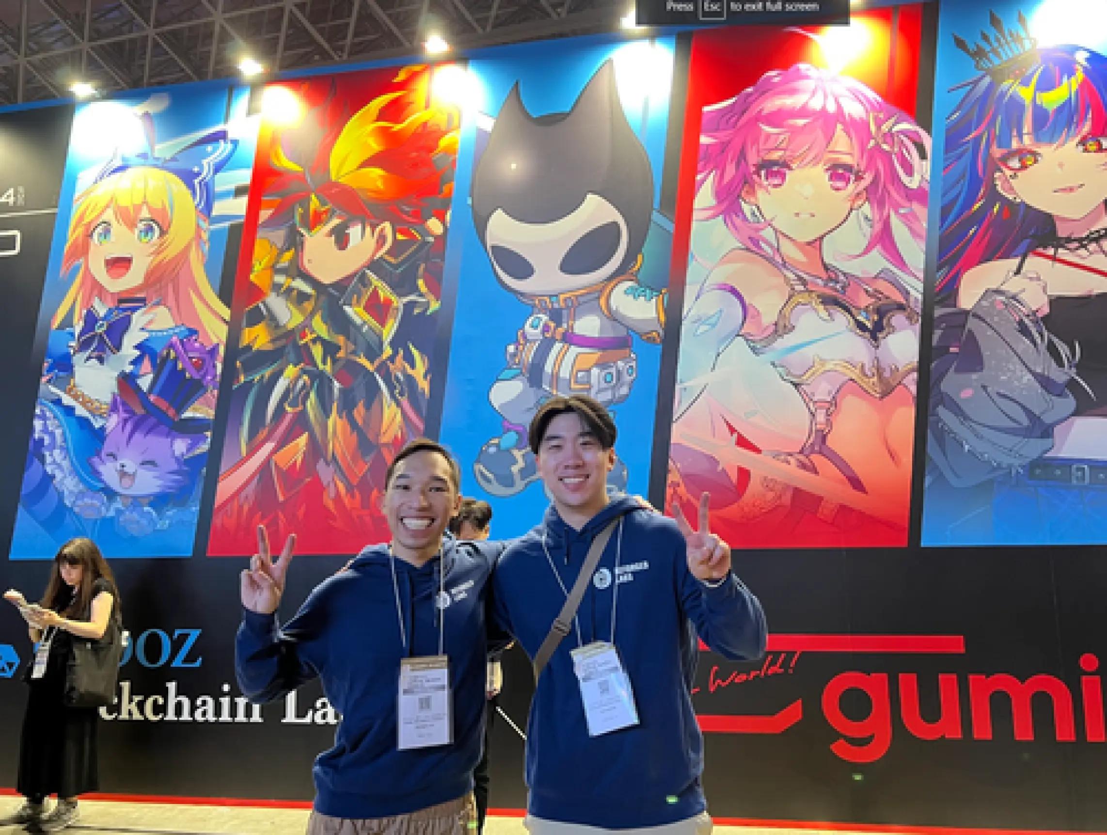 Robert Huynh and Oscar, CEO and CTO of Reforged Labs, smiling and posing in front of a vibrant anime-themed backdrop with colorful character illustrations at a convention or event.